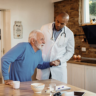 A healthcare professional assisting an elderly man at a table, with a cup and bowl visible in the setting.