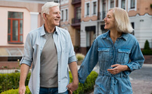 An older couple walking hand in hand, smiling at each other, dressed casually in a denim outfit and light shirt.