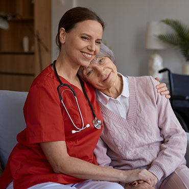 A caregiver in a red uniform with a stethoscope smiling while embracing an elderly woman in a pink sweater, both seated together.