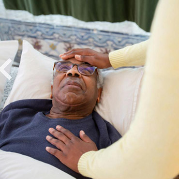 Elderly man lying in bed with a caregiver's hand on his forehead, showing a moment of care and attention in a home setting.