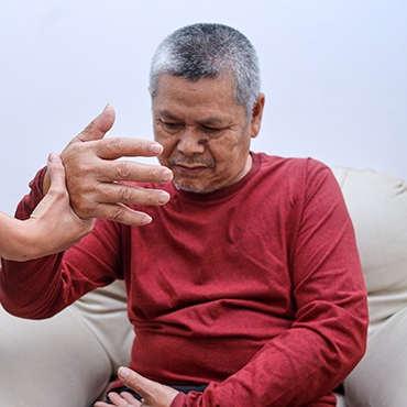 An elderly man in a red long-sleeve shirt sitting on a couch, with a hand reaching towards him, indicating a supportive interaction.