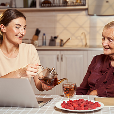 A caregiver pouring tea from a teapot into a glass for an elderly woman sitting at a table with a plate of raspberries.
