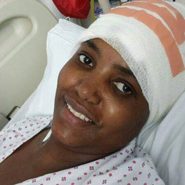 Patient with a bandaged head smiling while resting in a hospital bed, wearing a patterned hospital gown.