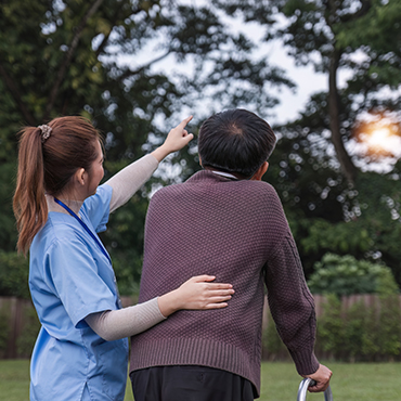 A caregiver assisting an elderly man in a sweater, as he points towards the sky while using a walker.