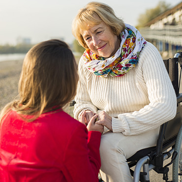 An elderly woman in a wheelchair wearing a colorful scarf, engaged in conversation with a younger woman in a red jacket.