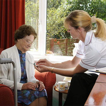 A caregiver assisting an elderly woman with her glasses while seated in a living room setting.