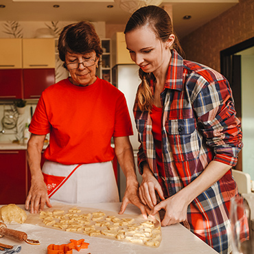 Two women preparing dough together in a kitchen, one wearing a red shirt and the other in a plaid shirt, focused on their task.